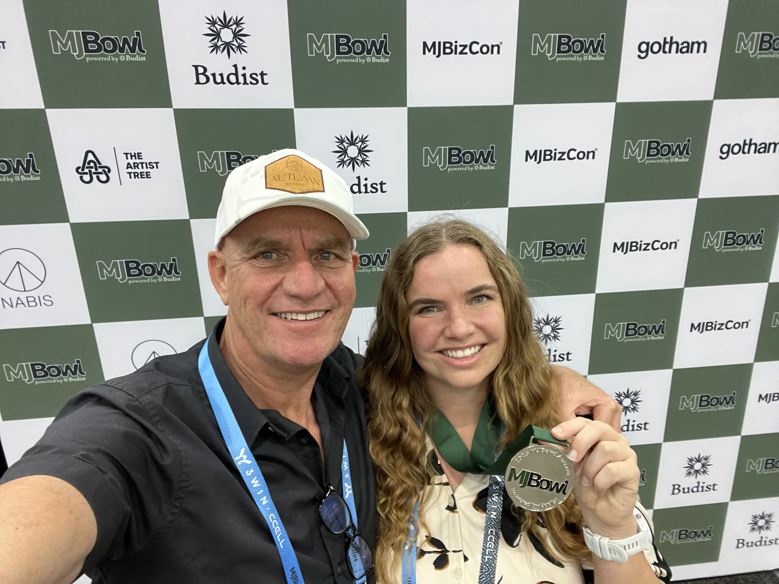 Hanna Brand and Hans Brand smiling together in front of an MJBizCon and MJBowl backdrop. Hanna is holding a silver MJBowl medal, and Hans has his arm around her as they take a selfie.